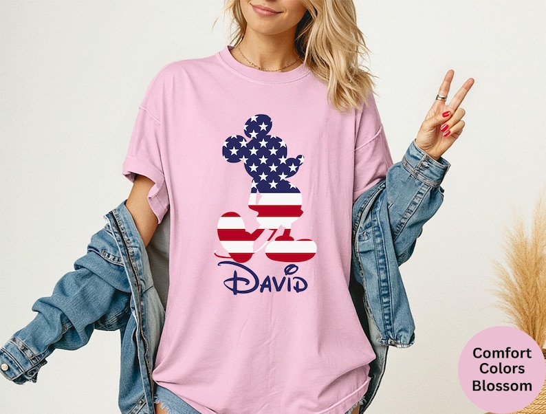 May include: A light pink t-shirt featuring a patriotic Mickey Mouse design. The Mickey Mouse silhouette is filled with the American flag pattern. The name "David" is written in a navy blue script font below the design.