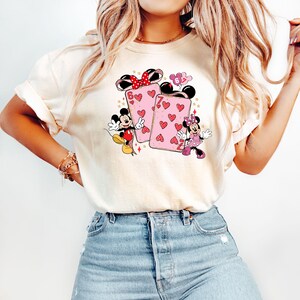 May include: Cream-colored t-shirt featuring a graphic of Mickey and Minnie Mouse with playing cards. The cards are pink with red hearts and the numbers 6 and 7. Mickey and Minnie are in their classic outfits.