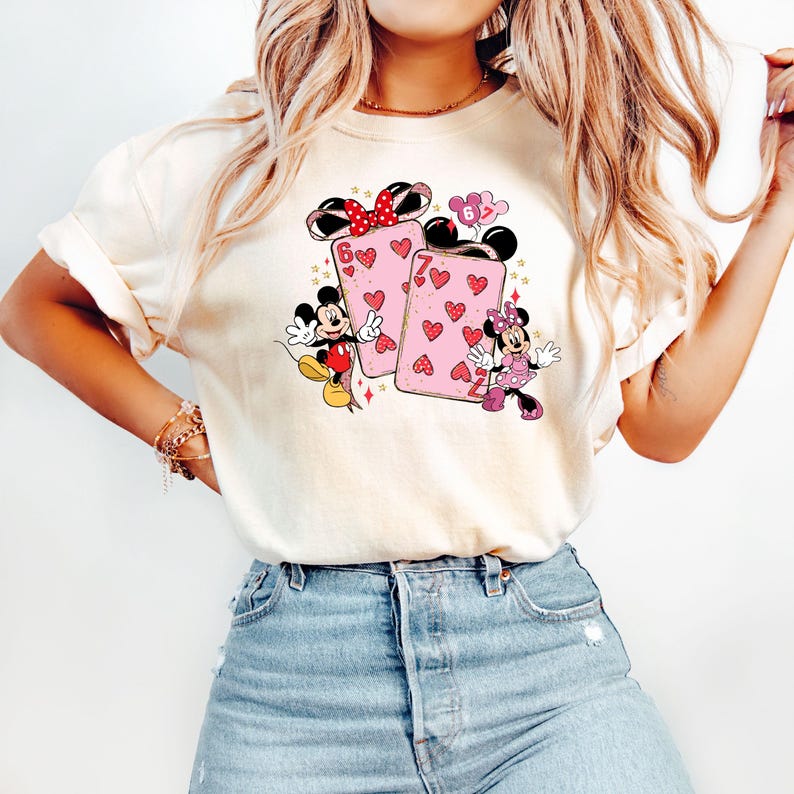 May include: Cream-colored t-shirt featuring a graphic of Mickey and Minnie Mouse with playing cards. The cards are pink with red hearts and the numbers 6 and 7. Mickey and Minnie are in their classic outfits.