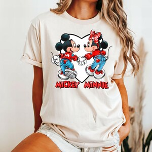 May include: A cream-colored t-shirt with a graphic of Mickey Mouse and Minnie Mouse holding hands inside a heart. The text "Mickey Minnie" is printed below the heart.
