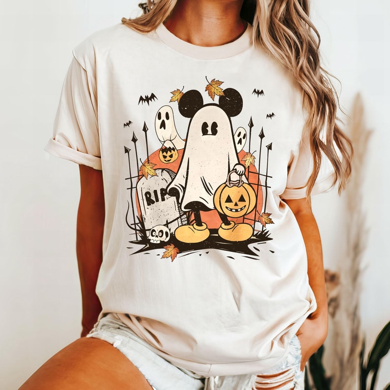 May include: A beige t-shirt with a cartoon image of Mickey Mouse dressed as a ghost holding a jack-o-lantern. The image includes a tombstone that says "RIP".