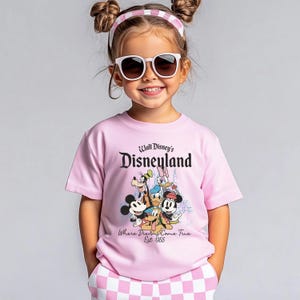 May include: A young person wearing a pink t-shirt with the text "Walt Disney's Disneyland" and a graphic of Disney characters. The shirt also has the text "Where Dreams Come True Est. 1955". The person is also wearing sunglasses and a pink and white headband.