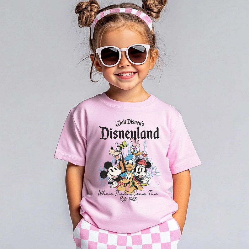 May include: A young person wearing a pink t-shirt with the text "Walt Disney's Disneyland" and a graphic of Disney characters. The shirt also has the text "Where Dreams Come True Est. 1955". The person is also wearing sunglasses and a pink and white headband.