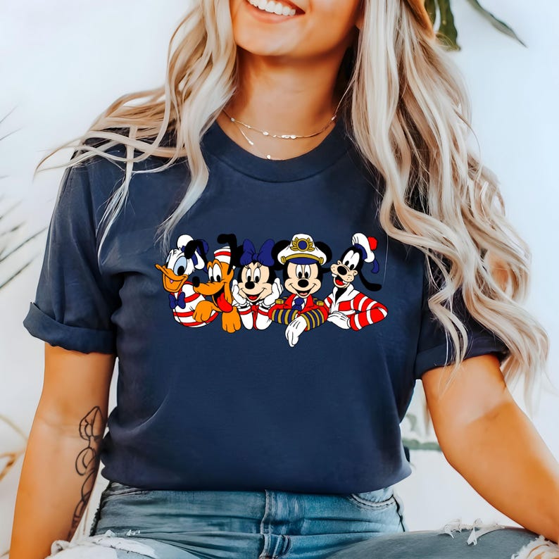Peut inclure: Un t-shirt bleu marine avec un motif de Mickey Mouse, Minnie Mouse, Donald Duck, Dingo et Pluto habill&eacute;s en marins. Les personnages sourient et saluent.