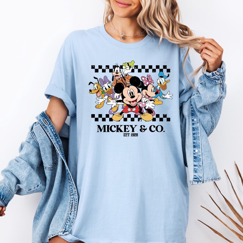 May include: Light blue t-shirt featuring a graphic of Mickey Mouse and friends, including Minnie Mouse, Donald Duck, and Goofy. The design is set against a black and white checkered pattern, with the text "Mickey & Co. Est 1928".