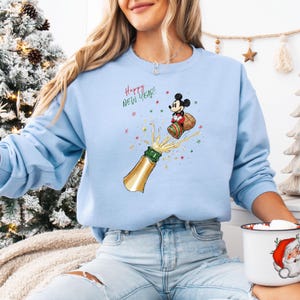 May include: Pale blue crew neck sweatshirt featuring a cartoon of Mickey Mouse celebrating the New Year with a champagne bottle. The words "Happy New Year!" are written above the graphic in red.