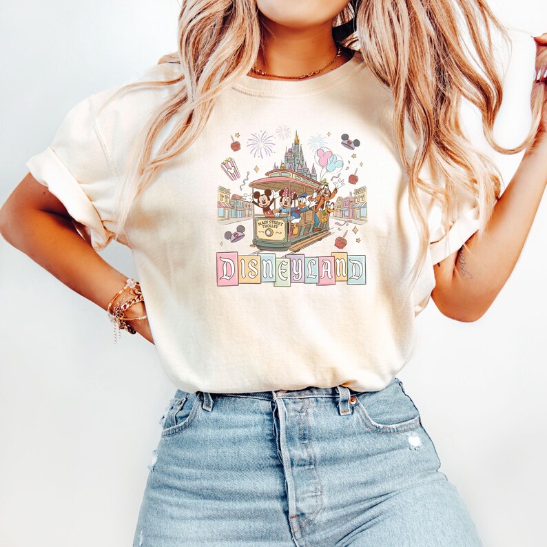 May include: Cream-colored t-shirt featuring a colorful Disneyland graphic. The design includes a trolley with Disney characters, a castle, fireworks, and the word "Disneyland" in pastel letters. The shirt is paired with blue jeans.