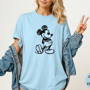 May include: Light blue t-shirt featuring a black and white sketch of Mickey Mouse. The shirt is a Comfort Colors Chambray and has a relaxed fit. The design is a classic cartoon character.