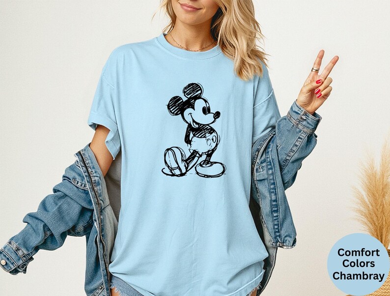 May include: Light blue t-shirt featuring a black and white sketch of Mickey Mouse. The shirt is a Comfort Colors Chambray and has a relaxed fit. The design is a classic cartoon character.