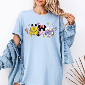 May include: Light blue t-shirt with a graphic of Mickey Mouse, Minnie Mouse, Pluto, and Daisy Duck spelling out the word "MOM".