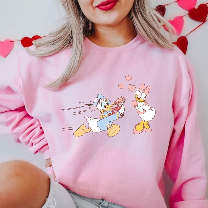 Op de afbeelding: Roze sweatshirt met een cartoon afbeelding van Donald Duck die met een bos bloemen naar Daisy Duck rent, die met gekruiste armen staat. Er zweven roze hartjes om hen heen.