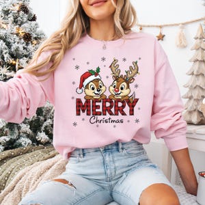 May include: A pale pink sweatshirt with a festive design. It features two cartoon chipmunks, one in a Santa hat and the other with reindeer antlers. The word "MERRY" is in a red plaid pattern, with "Christmas" below. Snowflakes are scattered around.