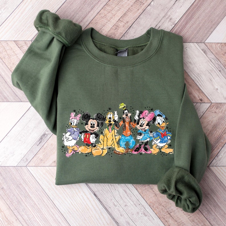 Puede incluir: Una sudadera verde con un estampado vintage de Mickey Mouse, Minnie Mouse, Donald Duck, Goofy, Pluto y Daisy Duck. Los personajes est&aacute;n todos sonriendo y de pie juntos.