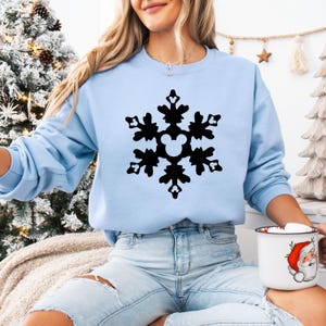 May include: A light blue sweatshirt featuring a large black snowflake design with a Mickey Mouse head in the center. The sweatshirt is a long-sleeved crew neck style. A white mug with a Santa Claus graphic is held in the hand.