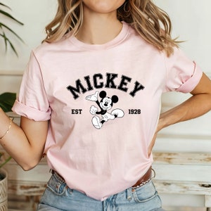May include: Pink t-shirt with a black and white graphic of Mickey Mouse. The graphic includes the text "Mickey" in a black arch above Mickey Mouse, "EST" to the left of Mickey Mouse, and "1928" to the right of Mickey Mouse.