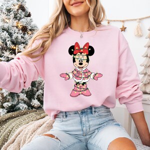 May include: A light pink sweatshirt featuring a graphic of Minnie Mouse in a pink winter coat and boots, adorned with Christmas lights. Minnie Mouse has a red bow and black ears. The sweatshirt is a solid light pink color.