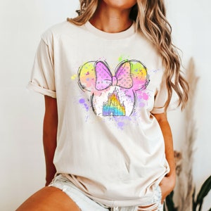 Puede incluir: Una camiseta beige claro con un gr&aacute;fico colorido de una cabeza de rat&oacute;n de dibujos animados con un lazo rosa y un castillo dentro de las orejas. El gr&aacute;fico est&aacute; rodeado de salpicaduras de pintura de colores.