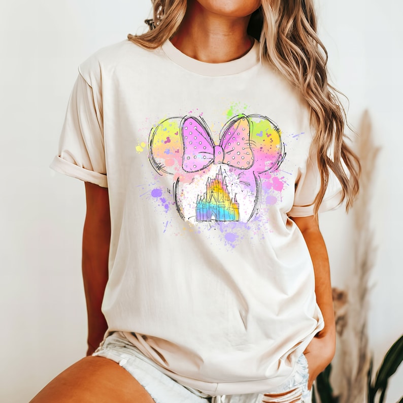 Puede incluir: Una camiseta beige claro con un gr&aacute;fico colorido de una cabeza de rat&oacute;n de dibujos animados con un lazo rosa y un castillo dentro de las orejas. El gr&aacute;fico est&aacute; rodeado de salpicaduras de pintura de colores.
