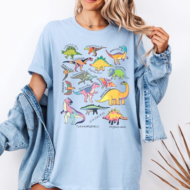May include: Light blue t-shirt with a colorful dinosaur graphic print. The dinosaurs are arranged in a circle and labeled with their names, including Stegosaurus, Triceratops, Tyrannosaurus Rex, and Spinosaurus.