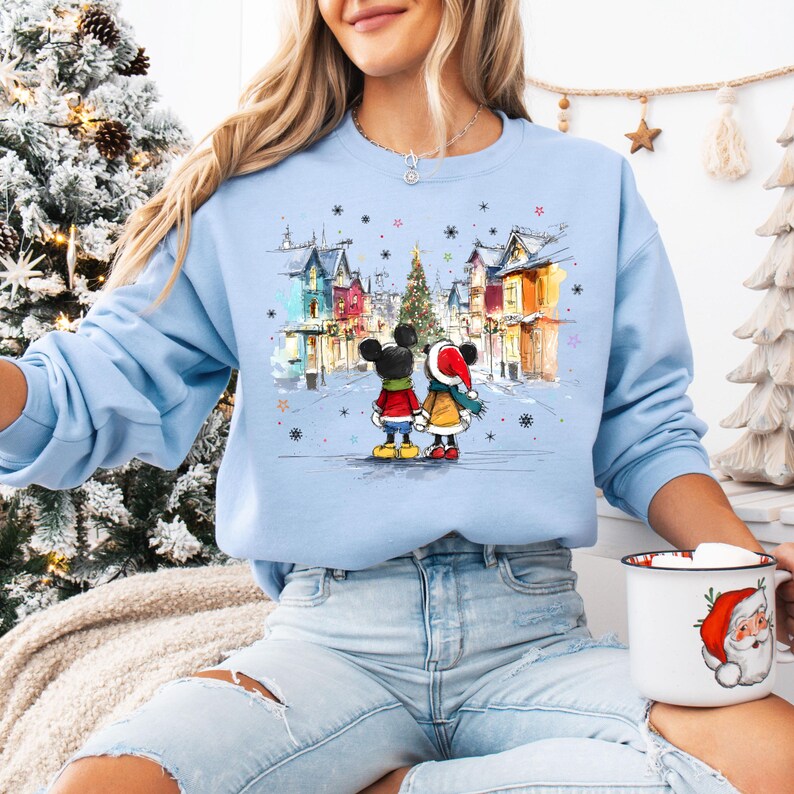 Op de afbeelding: Lichtblauw sweatshirt met een aquarel illustratie van Mickey en Minnie Mouse in een winterse sc&egrave;ne. Het ontwerp bevat een kerstboom, kleurrijke gebouwen en sneeuwvlokken. Het sweatshirt heeft een ronde hals.