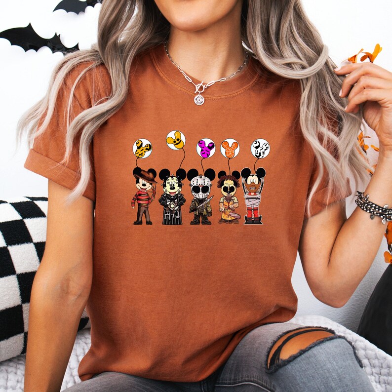 May include: Rust-colored t-shirt featuring a graphic of cartoon characters dressed as horror movie icons, each holding a balloon. The balloons are decorated with spooky patterns. The design is centered on the shirt.