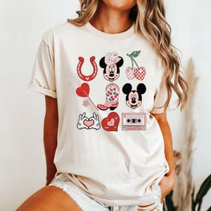 May include: A white t-shirt with a pink and red graphic design featuring a cartoon character wearing a cowboy hat, a horseshoe, a boot, a cherry, a heart, and a cassette tape.