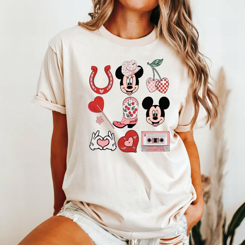 May include: A white t-shirt with a pink and red graphic design featuring a cartoon character wearing a cowboy hat, a horseshoe, a boot, a cherry, a heart, and a cassette tape.