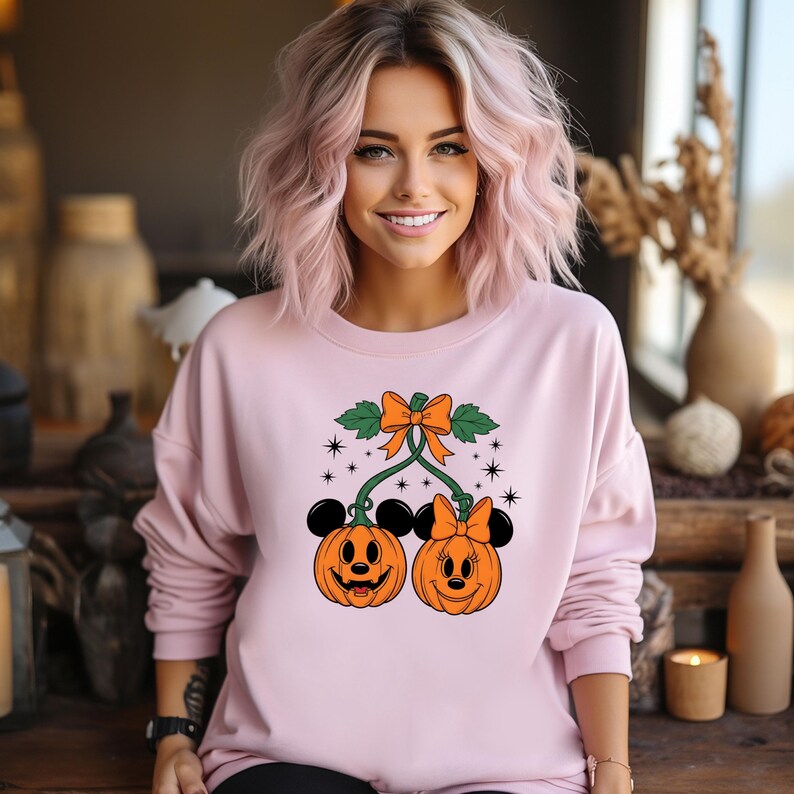 May include: A light pink sweatshirt featuring a Halloween-themed design. The graphic includes two orange jack-o'-lanterns with Mickey and Minnie Mouse ears, connected by green vines and an orange bow. Black stars and green leaves complete the design.
