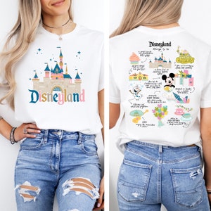 May include: A white t-shirt with a colourful illustration of Disneyland's Sleeping Beauty Castle on the front. The back of the shirt features a colourful illustration of various Disneyland attractions, including Mickey Mouse, churros, and the Frozen ride.