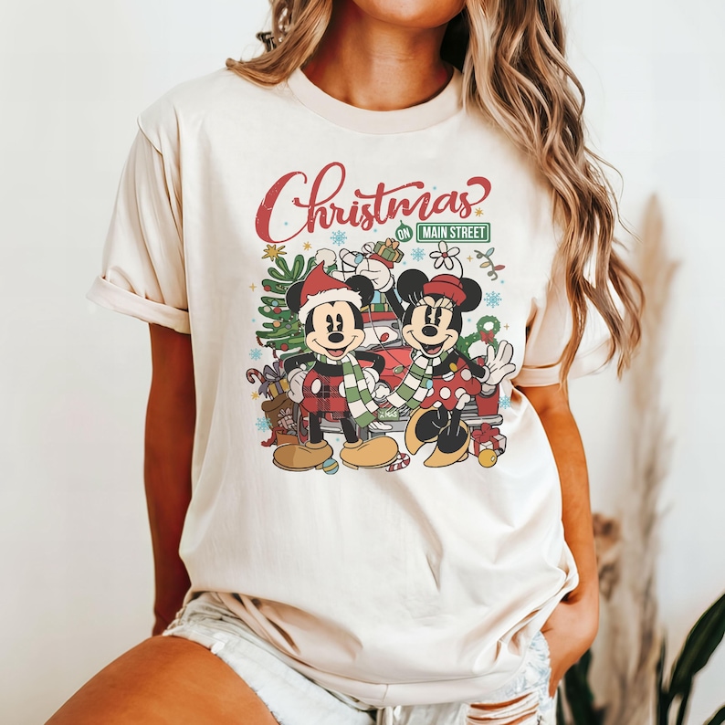 Op de afbeelding: Een cr&egrave;mekleurig T-shirt met een kerstthema met Mickey en Minnie Mouse. De afbeelding bevat de tekst "Christmas on Main Street".