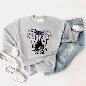 Puede incluir: Una sudadera gris claro con un gr&aacute;fico negro que presenta orejas de Mickey Mouse, un gorro de graduaci&oacute;n, fuegos artificiales y una silueta de castillo. El texto "Senior 2026" est&aacute; debajo del castillo. Zapatillas blancas y vaqueros azules tambi&eacute;n est&aacute;n en la imagen.
