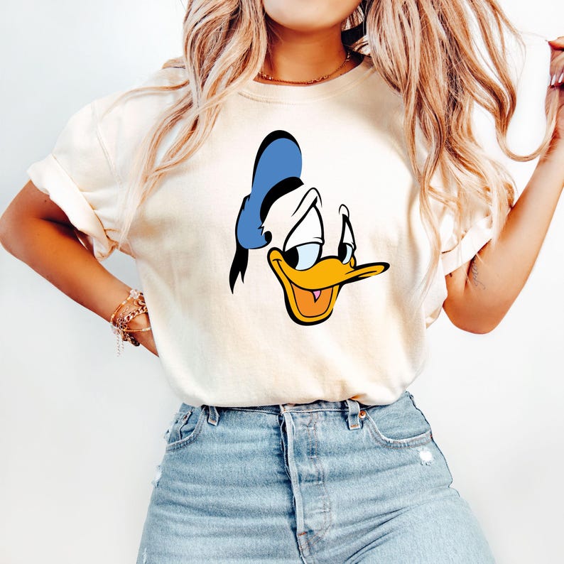 May include: Cream-colored t-shirt featuring a cartoon image of Donald Duck's face. The design includes a blue sailor hat, a yellow beak, and a smiling expression. The shirt is paired with blue denim jeans.