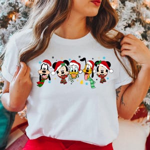 May include: White t-shirt with a Christmas-themed design featuring Mickey Mouse, Minnie Mouse, Donald Duck, Goofy, and Pluto wearing Santa hats and scarves.