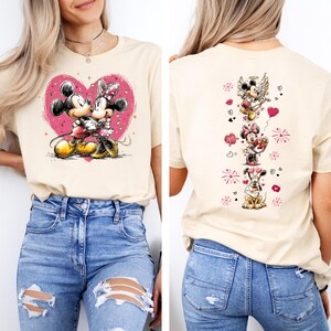 May include: A beige t-shirt featuring a graphic of Mickey and Minnie Mouse embracing within a pink heart. The back of the shirt displays a vertical design with various Disney characters and hearts. The shirt is paired with blue jeans.
