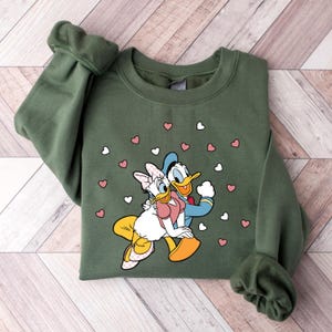 May include: A green sweatshirt with a cartoon image of Donald Duck and Daisy Duck holding hands. The image is surrounded by pink and white hearts.