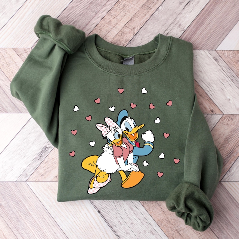 May include: A green sweatshirt with a cartoon image of Donald Duck and Daisy Duck holding hands. The image is surrounded by pink and white hearts.