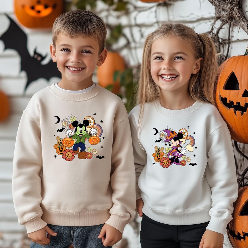 K&ouml;nnte beinhalten: Zwei Kinder tragen Halloween-Sweatshirts. Eines ist beige mit einem Mickey Mouse-Design, das K&uuml;rbisse h&auml;lt. Das andere ist wei&szlig; mit einem Minnie Mouse-Design in einem Hexenkost&uuml;m. K&uuml;rbisse und Flederm&auml;use sind im Hintergrund.