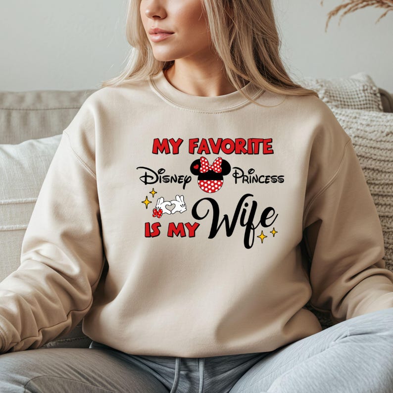 May include: A beige sweatshirt with the text "MY FAVORITE Disney Princess IS MY Wife" in red, black, and white. The design includes a Minnie Mouse bow and stars.