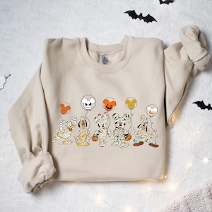 Peut inclure: Un sweat-shirt beige avec un motif de Mickey Mouse, Donald Duck, Dingo et Pluto habill&eacute;s en momies tenant des ballons. Les ballons sont en forme de t&ecirc;te de Mickey Mouse et sont de couleur orange, blanche et noire.