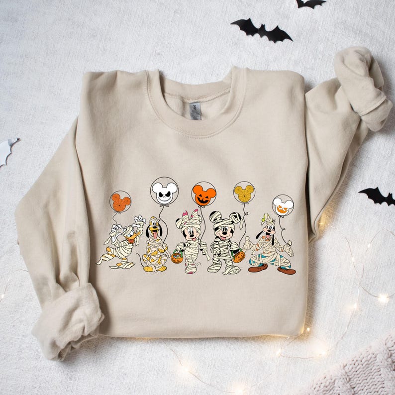 Peut inclure: Un sweat-shirt beige avec un motif de Mickey Mouse, Donald Duck, Dingo et Pluto habill&eacute;s en momies tenant des ballons. Les ballons sont en forme de t&ecirc;te de Mickey Mouse et sont de couleur orange, blanche et noire.