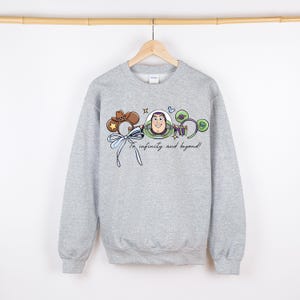 May include: A grey sweatshirt with a cartoon graphic of Buzz Lightyear and the phrase "To infinity and beyond!" in a script font. The design includes a cowboy hat, a bow, and other decorative elements. The sweatshirt is suspended from a wooden rail.