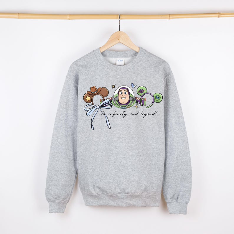 May include: A grey sweatshirt with a cartoon graphic of Buzz Lightyear and the phrase "To infinity and beyond!" in a script font. The design includes a cowboy hat, a bow, and other decorative elements. The sweatshirt is suspended from a wooden rail.