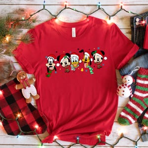 May include: Red t-shirt with a Christmas-themed design featuring Mickey Mouse, Minnie Mouse, Donald Duck, Goofy, and Pluto wearing Santa hats and scarves.
