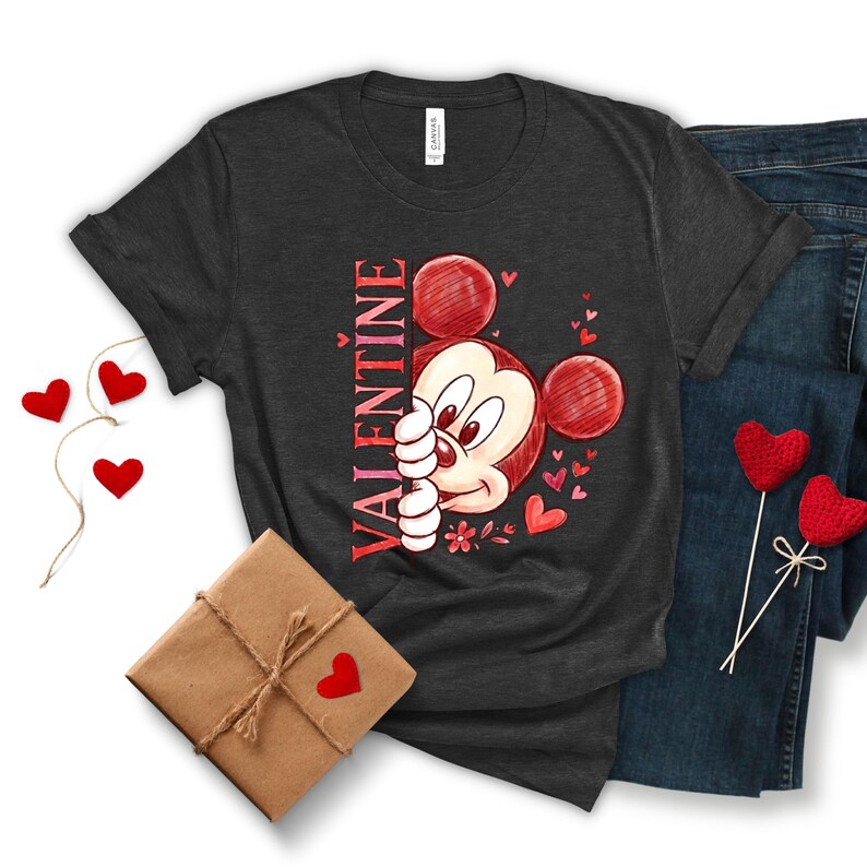May include: Dark grey t-shirt with a Valentine's Day design featuring Mickey Mouse. The word "VALENTINE" is printed vertically in red. The shirt is displayed with blue jeans, a wrapped gift, and heart decorations.