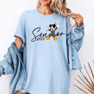 May include: Light blue t-shirt featuring a black and white graphic of Mickey Mouse wearing a graduation cap and gown. The text "Senior 2023" is printed in black below the graphic.