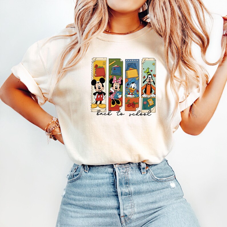 May include: Cream-colored t-shirt featuring a colorful graphic of Mickey Mouse, Minnie Mouse, Donald Duck, and Goofy with school supplies. The words "back to school" are printed below the characters. The shirt is made of a soft material.