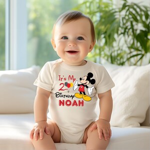 May include: A white bodysuit with a red and black Mickey Mouse graphic. The text "It's My 2nd Birthday NOAH" is printed on the bodysuit.