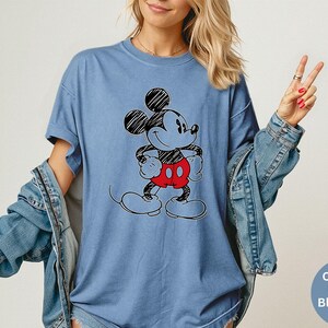 May include: A blue jean-colored t-shirt featuring a black and red Mickey Mouse graphic. The cartoon character is drawn with black outlines and has red shorts. The shirt has short sleeves and a relaxed fit.