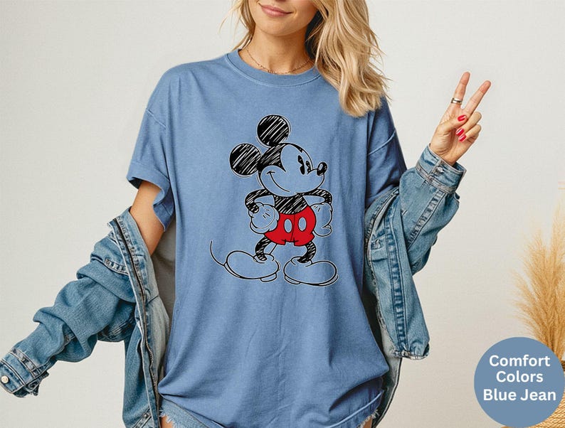 May include: A blue jean-colored t-shirt featuring a black and red Mickey Mouse graphic. The cartoon character is drawn with black outlines and has red shorts. The shirt has short sleeves and a relaxed fit.