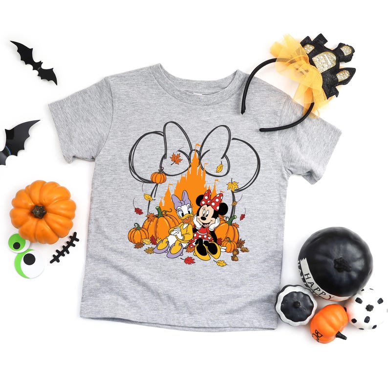 May include: A gray t-shirt featuring a Minnie Mouse and Daisy Duck graphic with pumpkins and autumn leaves. The design includes a large bow outline and a castle silhouette. Halloween-themed accessories surround the shirt.